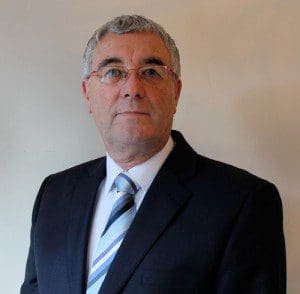 A man with short grey hair and glasses, exuding leadership, wears a dark suit, white shirt, and striped tie, standing against a plain light-coloured background.