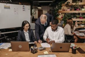 Three people in business attire work together at a desk with laptops and paperwork in an office. One person stands, pointing at a document, whilst the other two sit and look on. Plants and office supplies are in the background.