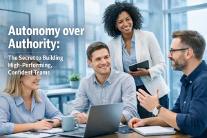 Four colleagues sit and stand around a table in a modern office, smiling and talking. The text on the image reads: “Autonomy over Authority: The Secret to Building High-Performing, Confident Teams”.