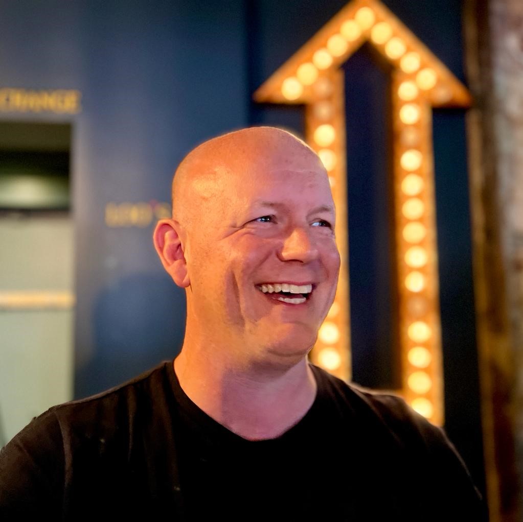Paul Bulloch smiling in a black shirt stands indoors, with a large illuminated arrow pointing upwards in the blurred background. The lighting is warm and the atmosphere appears cheerful.