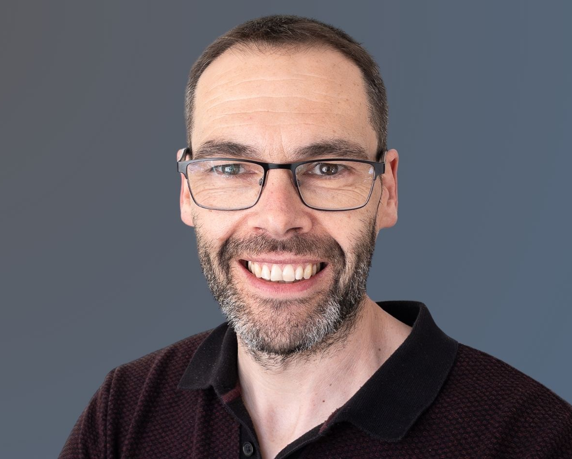A man with short brown hair, a beard, and glasses is smiling at the camera. He is wearing a dark collared shirt and is posed in front of a plain, gradient blue-grey background.