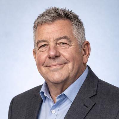 A middle-aged man with short grey hair, wearing a dark suit jacket and light blue collared shirt, smiles softly against a plain light blue background.
