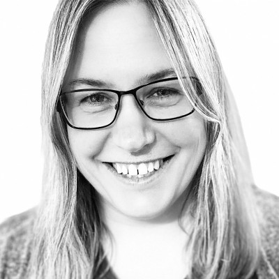 Laura Unwin profile picture -Smiling person with long, light-coloured hair and glasses, wearing a light top, shown in a black and white portrait against a plain background.