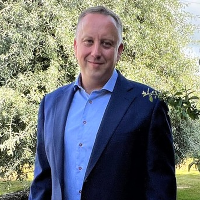 A man in a blue button-down shirt and a dark blazer standing outdoors in front of trees.